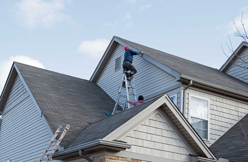 A roofing professional inspects a residential roof under partly cloudy skies with safety gear and measuring tools visible.